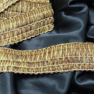 May include: Close-up of brown and gold fringe trim with a scalloped edge. The trim is on a black fabric background.