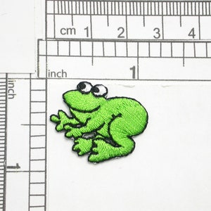 May include: Green embroidered frog applique patch with white eyes. The patch is approximately 1 inch wide.