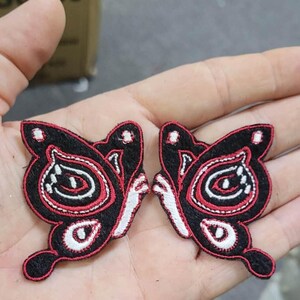 May include: Two embroidered butterfly patches featuring a black felt base with intricate stitching in red and white thread. The design includes bold red outlines, white accents, and a symmetrical pattern that mimics the shape of butterfly wings.
