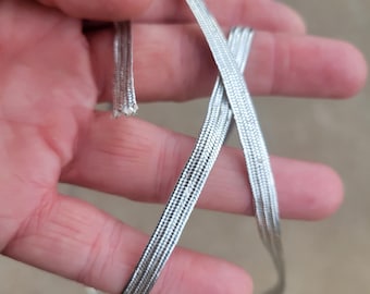 Puede incluir: Primer plano de un cordón trenzado plateado. El cordón está hecho de múltiples hebras entrelazadas, creando un diseño plano y flexible. El cordón se sostiene en una mano, con el extremo deshilachado. El cordón parece nuevo.