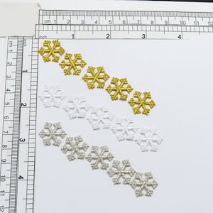 May include: A set of 10 snowflake embellishments in three colors: gold, silver, and white. The snowflakes are arranged in three rows, with the gold snowflakes at the top, the silver snowflakes in the middle, and the white snowflakes at the bottom.