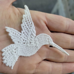 May include: A white, embroidered hummingbird applique with detailed wing and tail patterns. The delicate, openwork design features a long beak and is likely intended for fabric embellishment or craft projects.