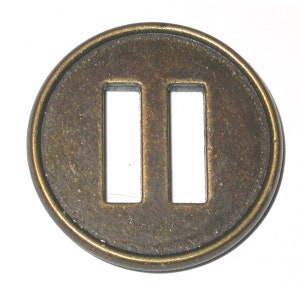 May include: A circular, antique bronze metal tsuba with two rectangular openings. The tsuba has a slightly textured surface and a dark, aged patina. The design is simple and symmetrical, with a raised outer rim.
