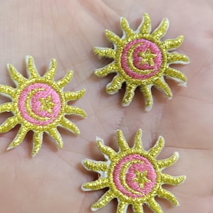 May include: Three circular embroidered patches featuring a central pink circle with a gold crescent moon and star design. Each patch is surrounded by radiating gold sun rays and has a white border, creating a sun-like celestial motif.