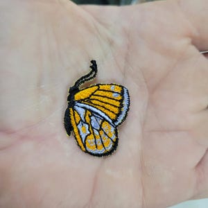 May include: Embroidered butterfly patch featuring yellow and black wings with blue and white accents. The patch has a detailed design and is likely for clothing or accessories.