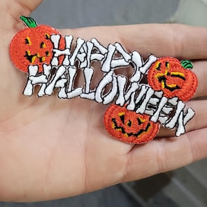 Pode incluir: Um emblema bordado com o texto HAPPY HALLOWEEN em letras brancas em forma de osso. O design inclui três abóboras de Halloween cor de laranja com caules verdes e traços faciais pretos esculpidos em torno dos elementos de texto.