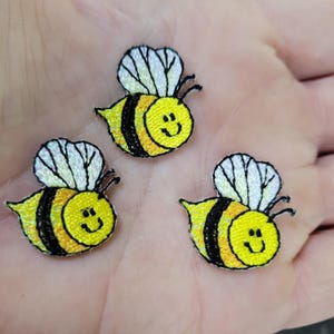 May include: Three embroidered bee patches. Each bee has a yellow body with black stripes, white iridescent wings, and a smiling face. The patches are small and likely for decorative purposes.