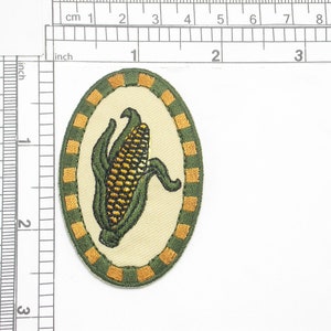 May include: An embroidered patch featuring a green and yellow corn cob on a cream background. The corn cob is surrounded by a green and gold border.