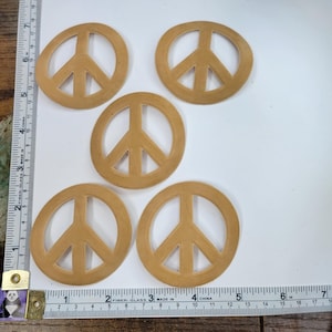 Peut inclure: Cinq découpes de signe de paix brun, chacune d'environ 5 cm de diamètre, sont disposées sur une surface en bois. Une règle est visible en bas de l'image.