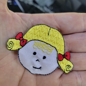 May include: Embroidered patch featuring a cartoon girl's face with yellow hair, red bows, and a smiling expression. The patch has a white face, black eyes, and a black outline. This patch is suitable for clothing or accessories.