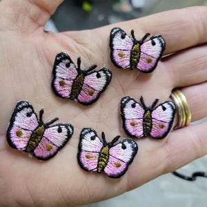 May include: Five embroidered butterfly patches. Each butterfly has pink wings with black outlines and a yellow and brown body. The patches are small and detailed, perfect for embellishing clothing or accessories.