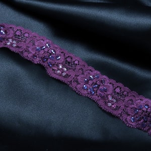 Pode incluir: Bordado de renda roxo com contas roxas iridescentes sobre um fundo de cetim preto.