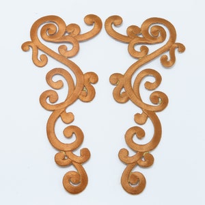May include: Two brown satin fabric appliques with a decorative scroll design. The appliques are symmetrical and have a delicate, intricate pattern.
