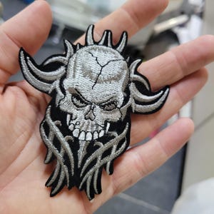 May include: Embroidered patch featuring a detailed skull design with horns and intricate silver and black stitching. The skull has a cracked forehead and a menacing expression, with stylized flames at the bottom. The patch is likely for jackets or bags.