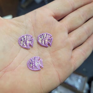 May include: Three small, embroidered fish appliques in shades of purple and iridescent silver are displayed. The fish have a detailed scale pattern and are outlined in a darker purple thread. The appliques are likely for crafting or embellishment.