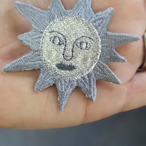 May include: A grey embroidered patch in the shape of a sun with a central face. The sun features a shimmering silver circular center with black stitched eyes, nose, and lips, surrounded by pointed grey rays radiating from the middle.