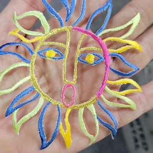 May include: An embroidered sun patch featuring a stylised face with a pink circular outline, yellow eyes, and a pink mouth. The sun rays are alternating in blue, lime green, and yellow thread, creating a vibrant, multi-coloured circular design.