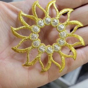 May include: A circular embroidered patch featuring a sun-like design with golden yellow rays extending outward from a center ring of twelve small, metallic silver circular accents. The embroidery has a raised, textured finish throughout.