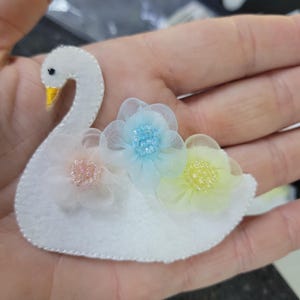 May include: A white felt swan applique with a yellow beak and black eye. The swan is decorated with three pastel-colored fabric flowers: pink, light blue, and yellow. The applique is designed for crafts and embellishments.