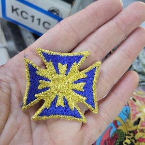May include: Embroidered patch featuring a blue and gold cross design. The patch has a gold center with a starburst pattern, surrounded by a blue border. The patch is held in a hand.