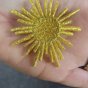Op de afbeelding: Een cirkelvormige gouden geborduurde patch met een zonnestraalontwerp met talloze uitstralende stralen van verschillende lengtes. De centrale schijf en de omliggende punten zijn gemaakt van metallic gouden draad met een textuur.
