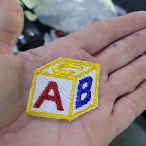 May include: A fabric patch in the shape of a child's alphabet block. The block is white with yellow trim and features the letters A, B, and C in red and blue. The patch is held in a hand.