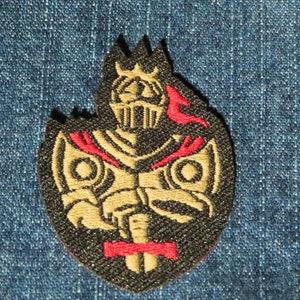 May include: A black and gold embroidered patch of a knight in armor holding a sword. The knight is wearing a red cape and a crown. The patch is on a blue denim background.