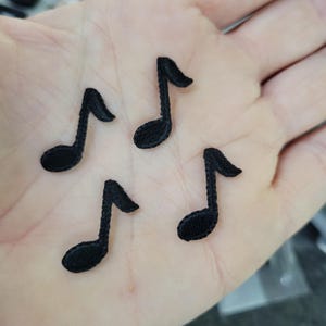Op de afbeelding: Vier kleine, zwart geborduurde muzieknoot patches. De patches hebben de vorm van achtste noten, met een gebogen steel en een afgeronde nootkop. De patches zijn waarschijnlijk bedoeld om op stof te naaien of te strijken.