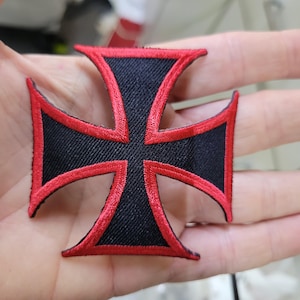 May include: Embroidered black cross patch with red trim. The patch features a four-armed cross design with pointed ends. The red trim outlines the edges of the cross, creating a bold contrast against the black fabric. This patch is suitable for jackets, bags, or other fabric items.