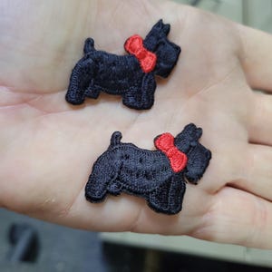 May include: Two black embroidered dog patches with red bowties. The patches are in the shape of a Scottish Terrier. The patches are small and detailed, with visible stitching. The patches are likely for sewing or crafting.