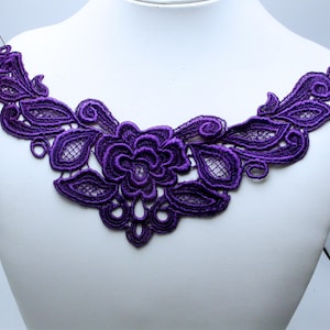 Puede incluir: Una aplicación de encaje de color morado intenso con un motivo floral central rodeado de patrones simétricos de hojas y enredaderas. El intrincado bordado incluye detalles de malla calada y está sujeto en los extremos con dos alfileres blancos.