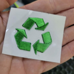 May include: A green embroidered recycling symbol on a white square. The symbol features three arrows in a circular pattern, representing the concept of reduce, reuse, and recycle. The design is a patch or sticker.