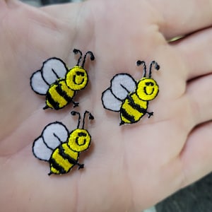 May include: Three embroidered bee patches. Each bee has a yellow body with black stripes, white wings, and black antennae. The bees are smiling. These patches are suitable for clothing or craft projects.