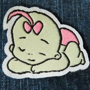 May include: A white embroidered patch of a sleeping baby with a pink bow and a pink diaper. The baby is outlined in black and has a light green skin tone.