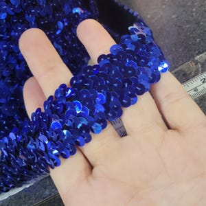 May include: Close-up of a blue sequined trim. The trim is made of small, round sequins that are sewn together in a continuous pattern. The sequins are a vibrant blue color and have a slight iridescent sheen.