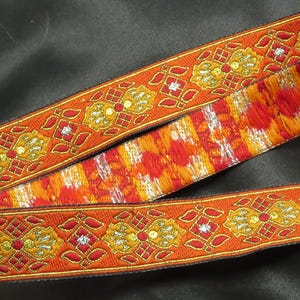 May include: Orange and gold embroidered ribbon with a repeating floral pattern. The ribbon has a textured, fringed section with red, orange, and yellow hues. The ribbon is likely used for crafting or embellishment.