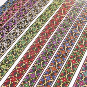 Pode incluir: Conjunto de fitas tecidas coloridas com fundo preto e detalhes dourados. A fita apresenta um padrão geométrico repetitivo com desenhos em forma de estrela.