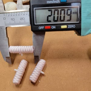 May include: A digital caliper measures a light pink, cylindrical object at 20.09 mm. Two additional pink cylinders with white plastic attachments are also visible. The objects are on a brown surface.