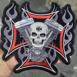 Op de afbeelding: Geborduurde patch met een schedel met een kruis op het voorhoofd, tegen een rood en zwart kruisontwerp. Het ontwerp bevat motoronderdelen en grijze vlammen, met een zwarte rand.