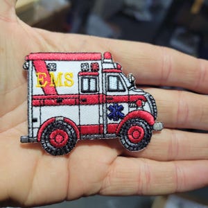 Può includere: Toppa ricamata di un'ambulanza rossa e bianca con ruote nere. La toppa presenta le lettere "EMS" in giallo e un emblema blu a forma di stella della vita. Il design dell'ambulanza include finestre e luci di emergenza.