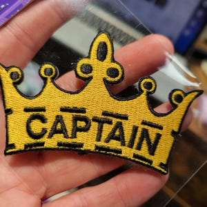 Peut inclure: Un écusson brodé jaune avec un contour noir d'une couronne et le mot "CAPTAIN" en lettres noires.