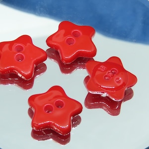 May include: Four glossy red star-shaped buttons with two holes each. The buttons are arranged on a reflective surface, creating a mirrored effect. The buttons are a vibrant red color.