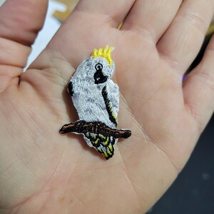 May include: Embroidered patch featuring a white cockatoo bird with a yellow crest and black details, perched on a brown branch. The patch is designed with various colours and textures, ideal for clothing or accessories.