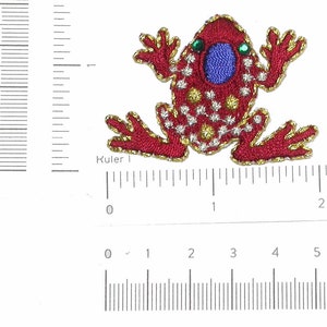 Pode incluir: Aplique de sapo bordado em vermelho com acabamento dourado e olhos azuis. O sapo tem aproximadamente 2,5 cm de largura.