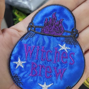 Op de afbeelding: Een blauwe geborduurde patch in de vorm van een ketel met een zwarte rand. De patch heeft paarse vlammen aan de bovenkant, de paarse tekst "Witches Brew" in het midden en drie witte geborduurde sterren verspreid over het blauwe oppervlak.