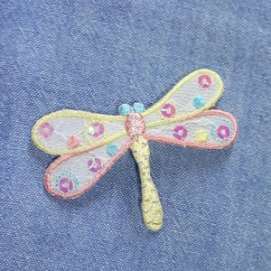 May include: A white, pink, and yellow embroidered dragonfly with colorful sequins. The dragonfly is on a blue denim background.