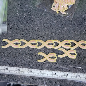 Könnte beinhalten: Hellrosa und goldfarbene Bandverzierung mit einem Unendlichkeits-Symbol-Design. Mehrere Teile sind sichtbar, darunter ein langer Streifen und einzelne Symbole. Ein Lineal mit cm-Maßen ist im Vordergrund.