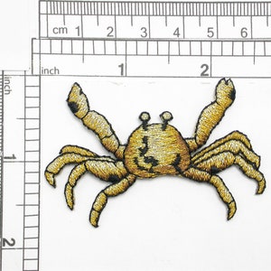 May include: A gold embroidered iron-on patch of a crab with black eyes. The crab is about 2 inches wide.