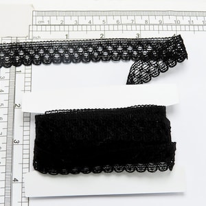 May include: Two pieces of black lace trim, one is a narrow trim with a scalloped edge and the other is a wider trim with a more intricate design. The trim is measured against a ruler in inches.