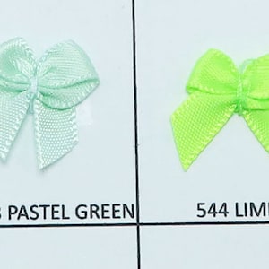 May include: A selection of ten small satin bows in various colors, including ultraviolet, amethyst, grappa, pastel green, lime, apple, greenflash, and emerald. The bows are arranged in a row on a white background.